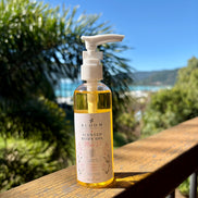 Miss D scented body oil 100 mL with white pump on a timber railing with tropical greenery and a coastal view