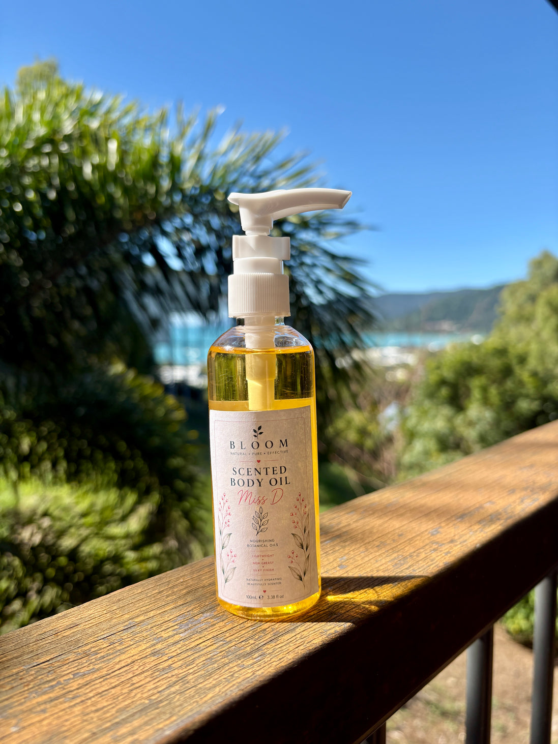 Miss D scented body oil 100 mL with white pump on a timber railing with tropical greenery and a coastal view