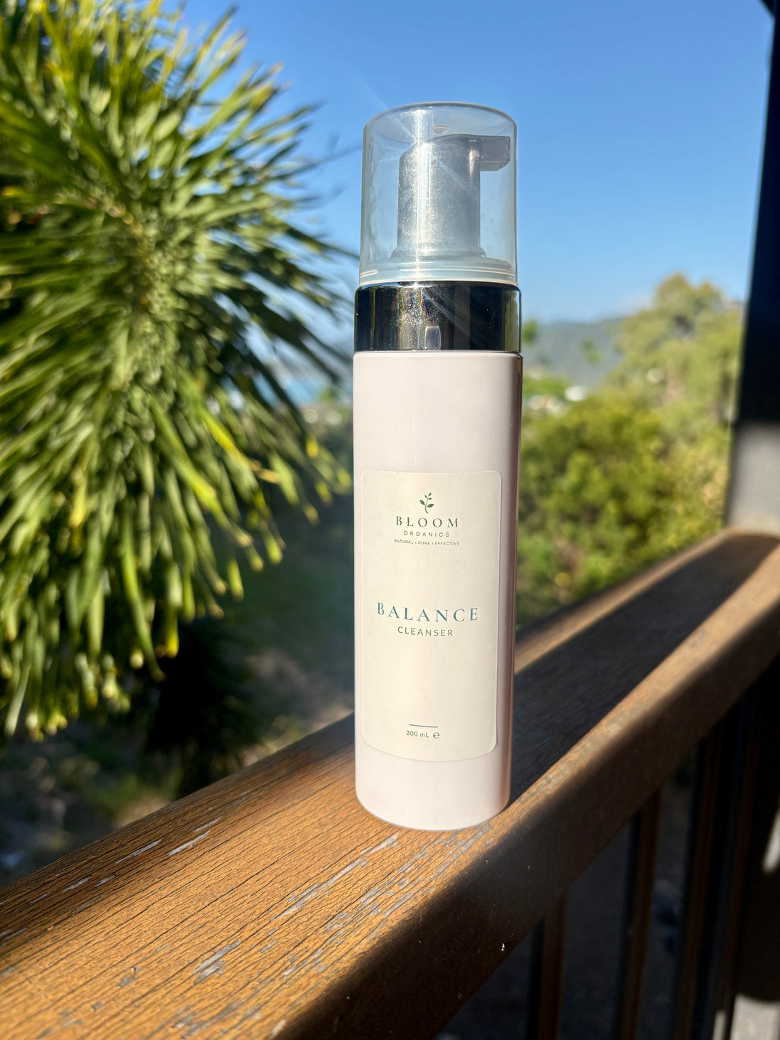 Bloom Organics Balance Cleanser, 200 mL pump bottle on a wooden railing with greenery behind it.