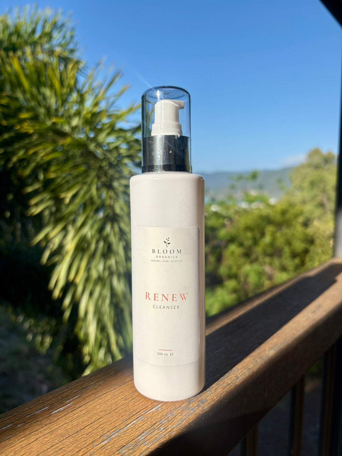 Bloom Organics Renew Cleanser, 200 mL pump bottle on a wooden railing with greenery behind it.