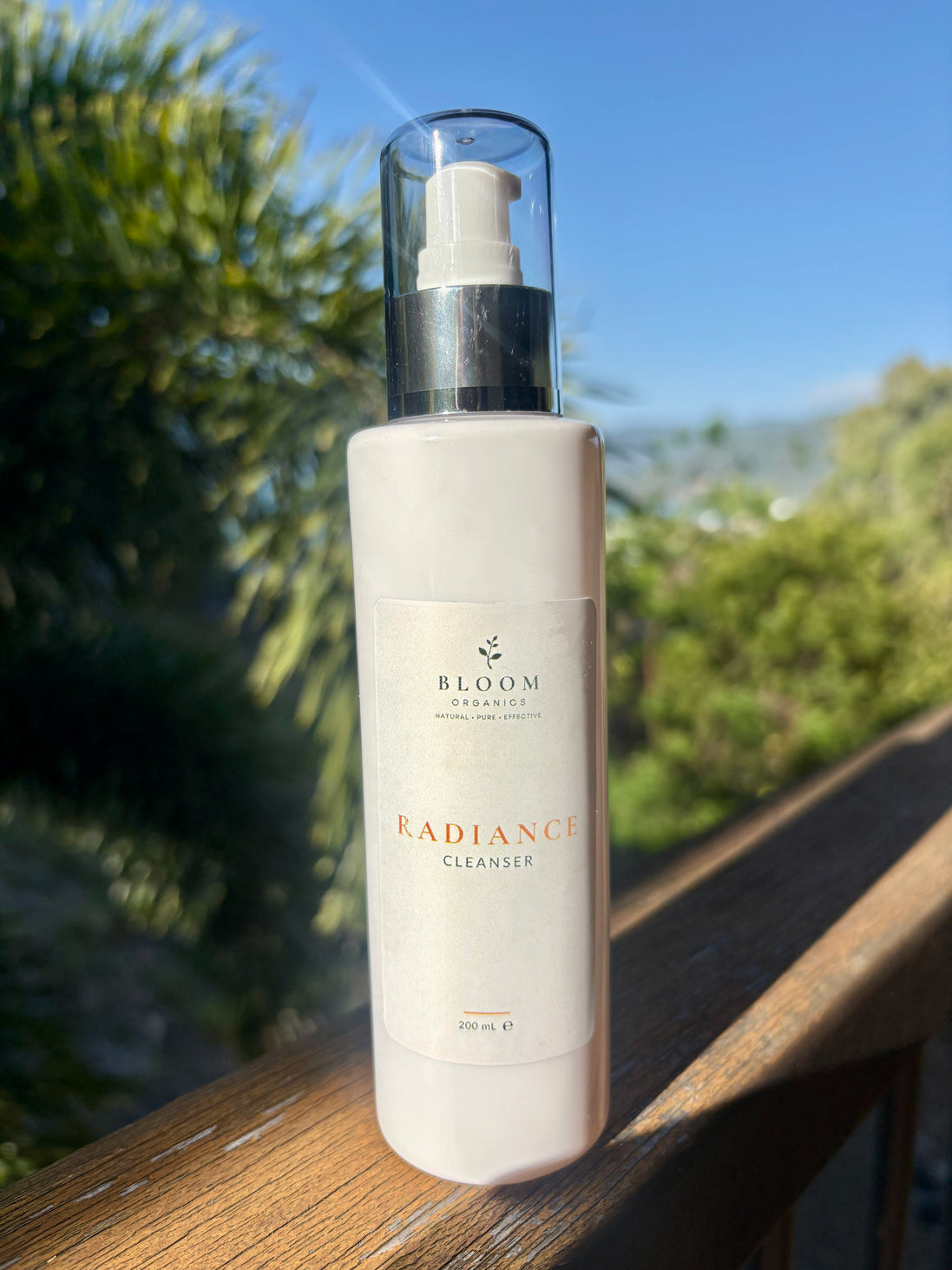 Bloom Organics Radiance Cleanser, 200 mL pump bottle on a wooden railing with greenery behind it.