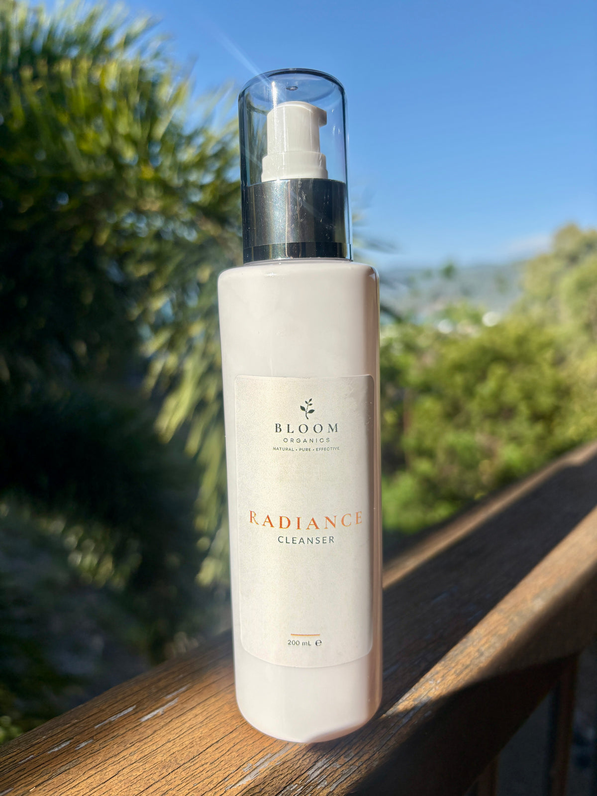 Bloom Organics Radiance Cleanser, 200 mL pump bottle on a wooden railing with greenery behind it.
