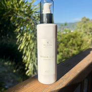 Bloom Organics Hydrate Cleanser, 200 mL pump bottle on a wooden railing with greenery behind it.