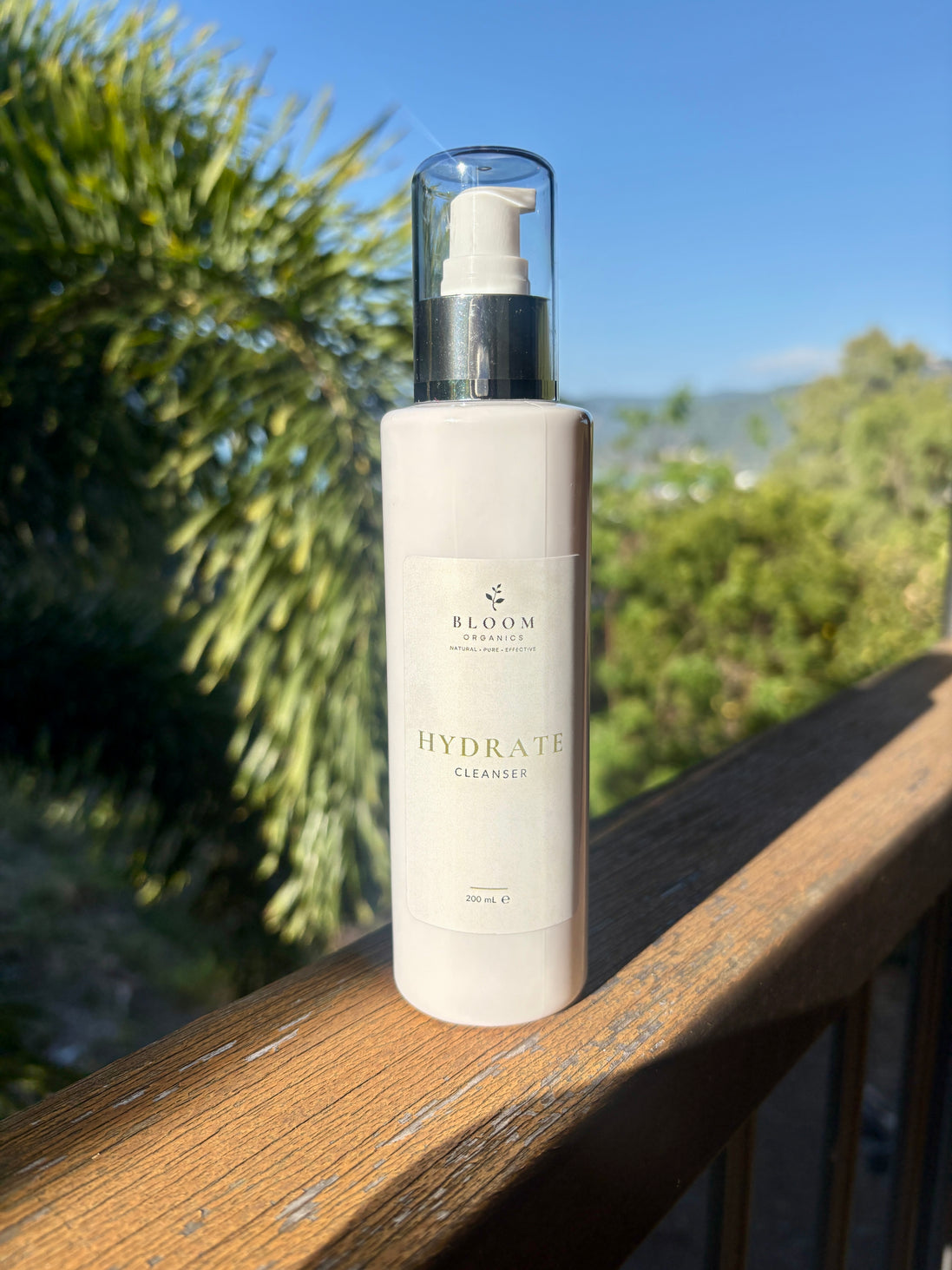 Bloom Organics Hydrate Cleanser, 200 mL pump bottle on a wooden railing with greenery behind it.